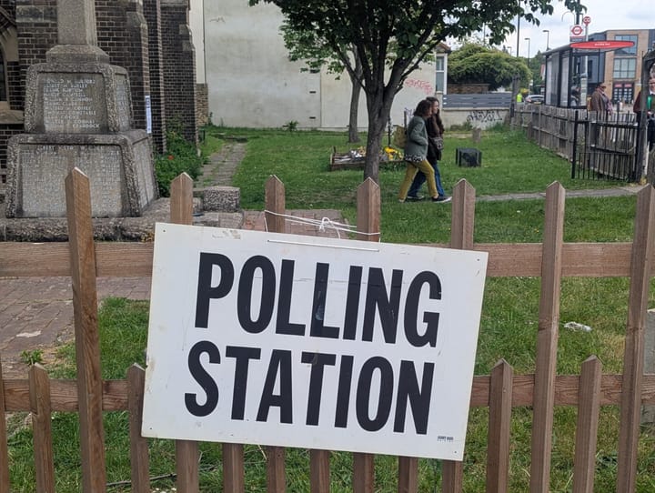 Notice on wooden fence saying Polling Station, church yard behind. 