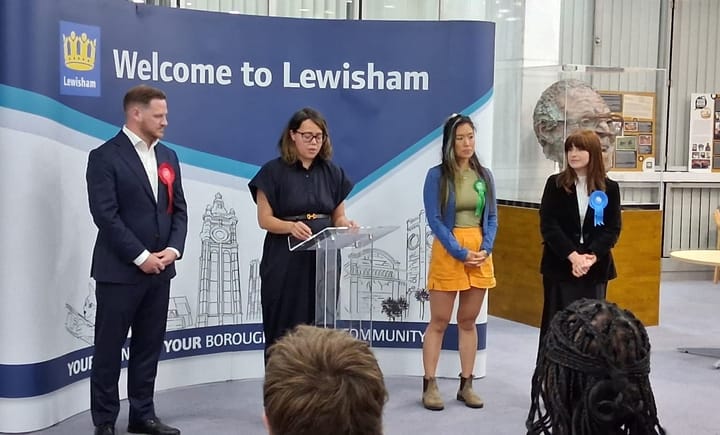 Candidates wearing party rosettes standing around woman at a lectern reading results.