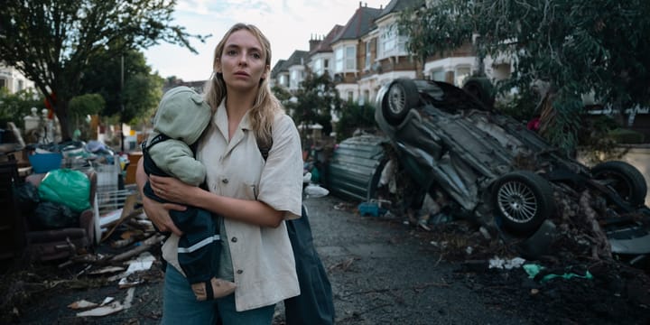 Woman carrying a baby in her arms, in a suburban street - household contents piled at either side and overturned cars. 