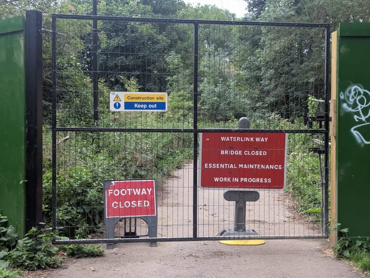 High Iron gate locked across the path with signs behind it, footway close, bridge closed, essential maintenance.
