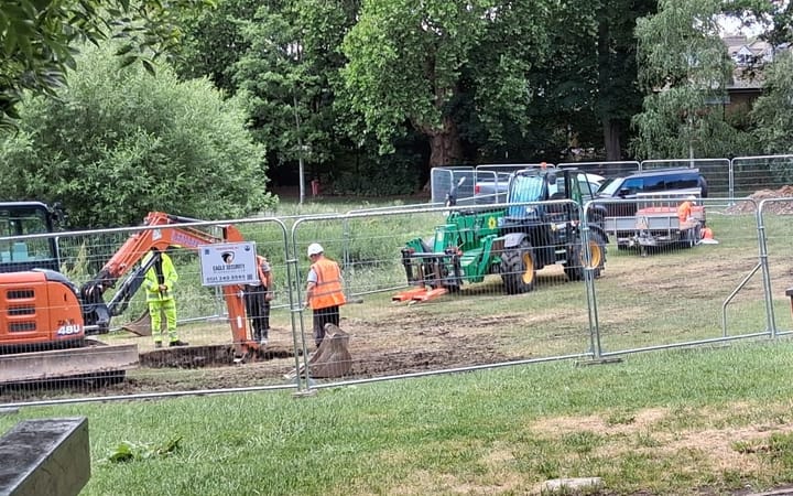 Workers in high vis and heavy machinery and diggers behind fencing in the park.