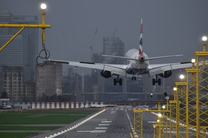 Plane landing at London City Airport under stormy sky, tower blocks and HSBC building in background.