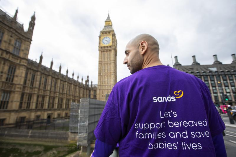 Campaigning parent from Sands in sweatshirt reading "let's support bereaved families and save babies' lives", Westminster and Big Ben in the background. at Westminster earlier this year
