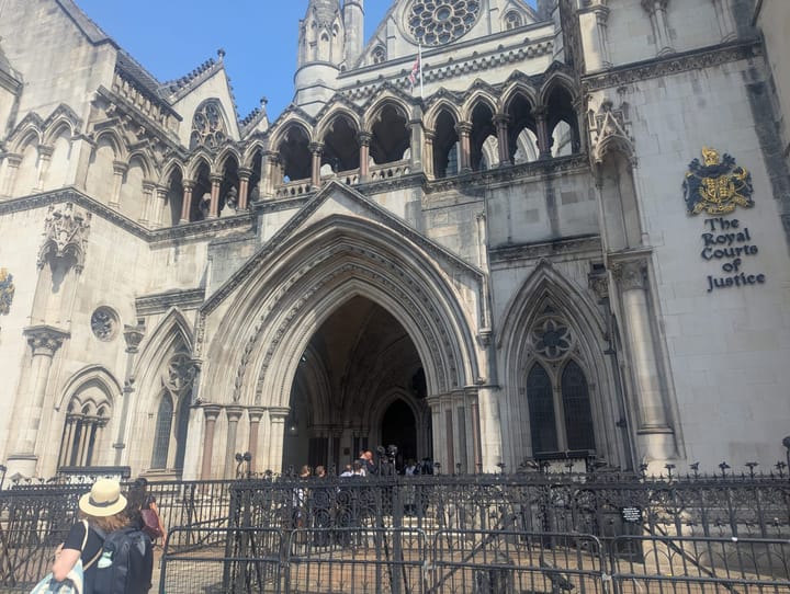 People queuing and passersby in straw hats outside the entrance to the gothic Royal Courts of Justice on The Strand.