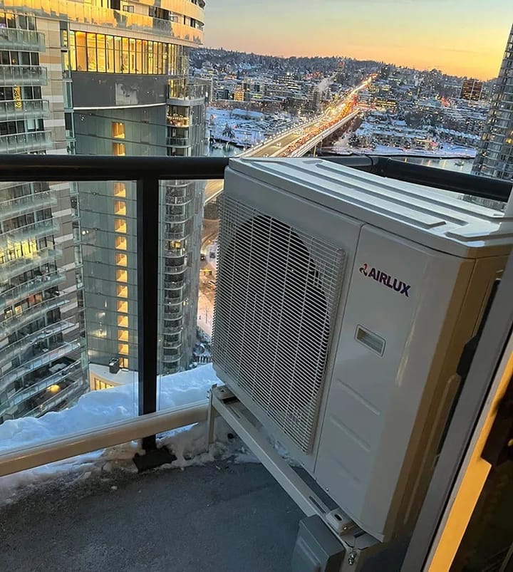 Outdoor heat pump on a balcony, snow visible at edge of balcony, view over Vancouver at sunset.t.