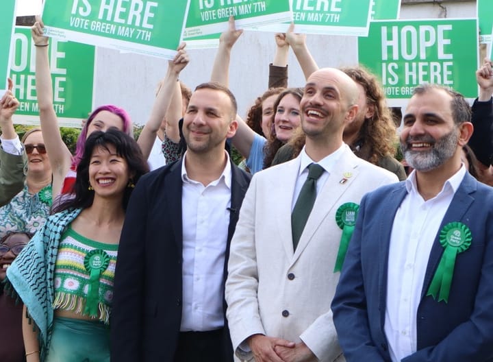  Hau-Yu Tam, Zack Polanski, Liam Shrivastava and Tauseef Anwar, smiling and wearing rosettes after the local election mayoral count.ts. 