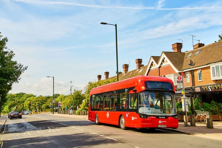 Single decker red bus at stop outside a shopping parade, sunlit street. 