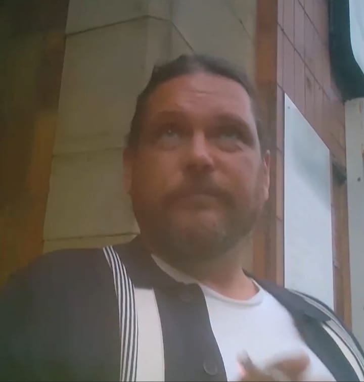 Screen grab from the filming, bearded man in white T shirt and dark blue and white light sports jacket, looking up and not aware of camera.